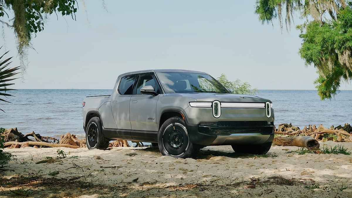 A gray Rivian R1T electric pickup parked on sand near water and trees.