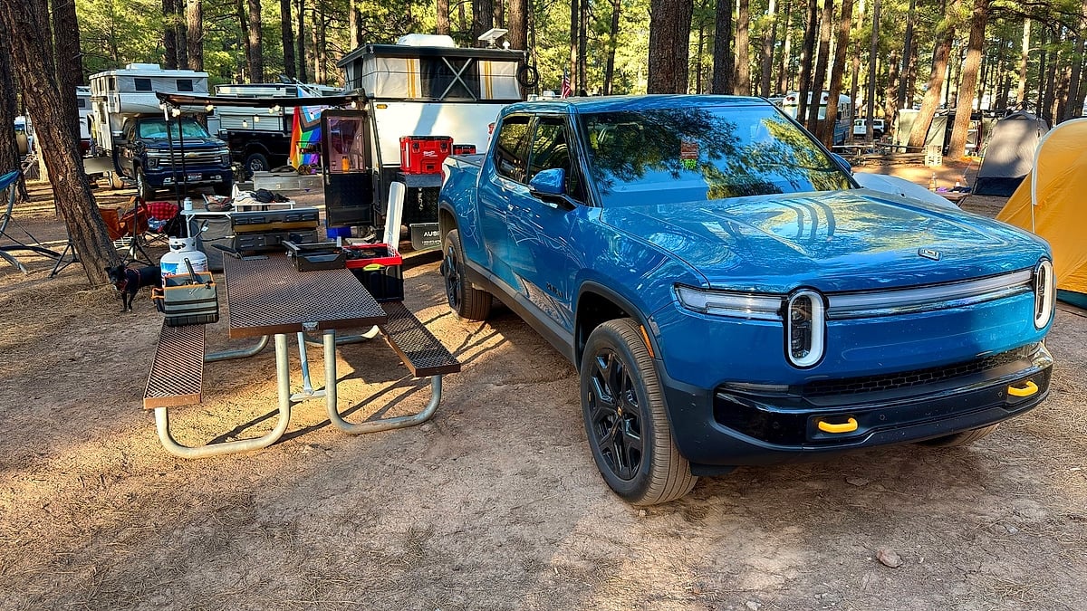 Blue Rivian R1T parked at a forest campsite with an off-road camper trailer, picnic table, and camping gear.