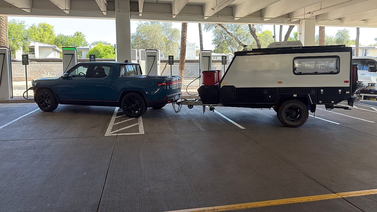 Blue Rivian R1T towing an off-road camper trailer at a covered EV charging station.