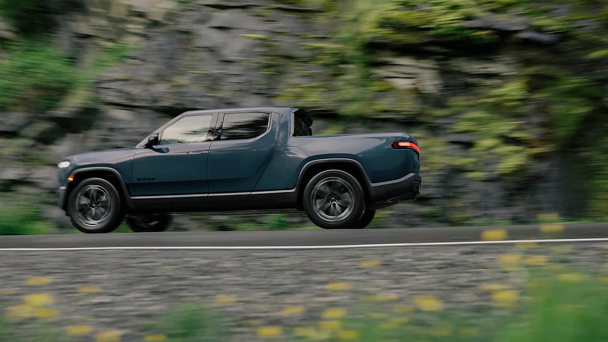 Rivian R1T electric pickup driving along a road beside a mossy rock wall with motion blur.