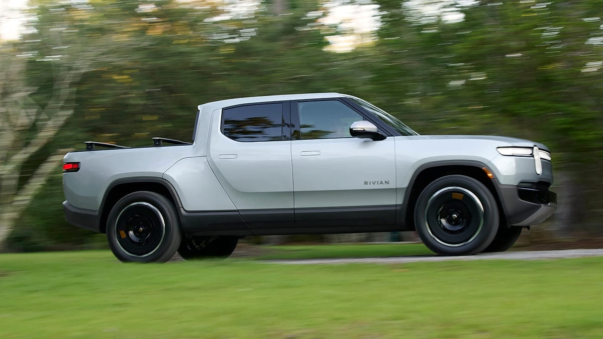 A silver Rivian R1T electric pickup drives past blurred green trees.