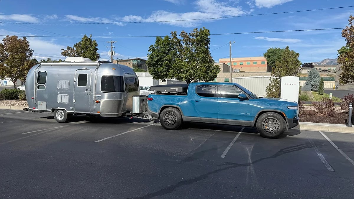 Blue Rivian R1T towing an Airstream travel trailer parked near a Tesla Supercharger in a parking lot.