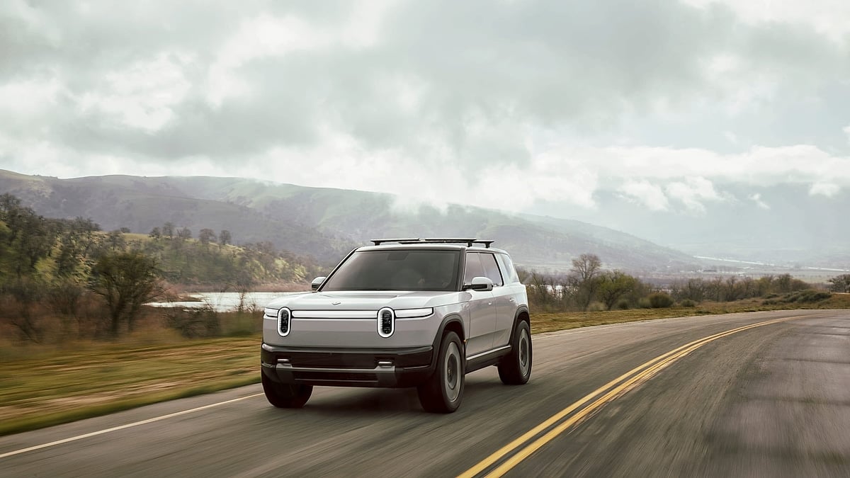 White Rivian R2 driving on an open road beneath dramatic clouds with hills and water in the distance.