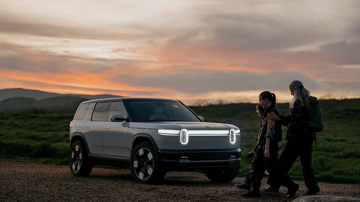 2027 Rivian R2 electric SUV parked at sunset with headlights illuminated as a family walks toward it on a gravel hillside
