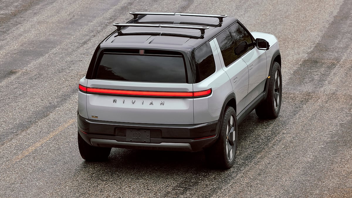 Silver Rivian R2S shown from a rear three-quarter angle on a paved road with roof crossbars.