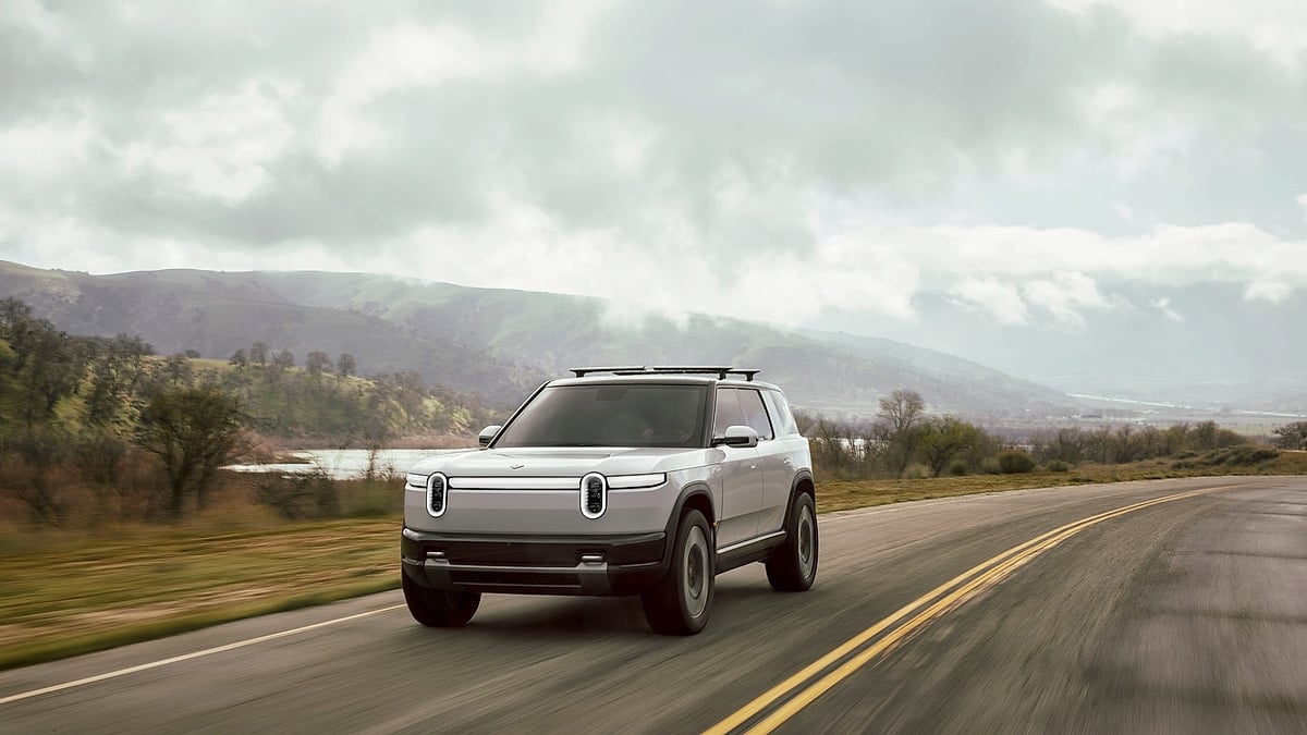 Silver Rivian R2S driving on a two-lane road with cloudy hills and water in the background.