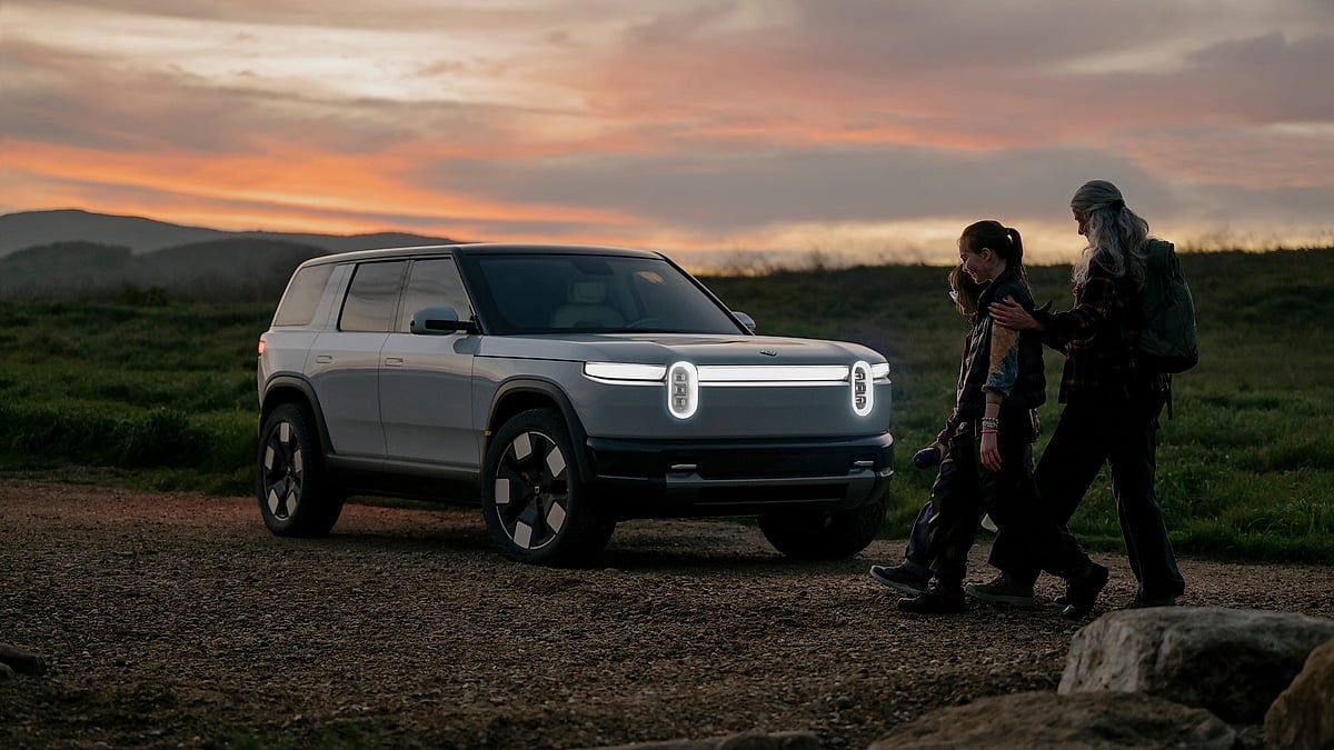 White Rivian R2 parked at sunset as two hikers walk beside it on a rural trail.