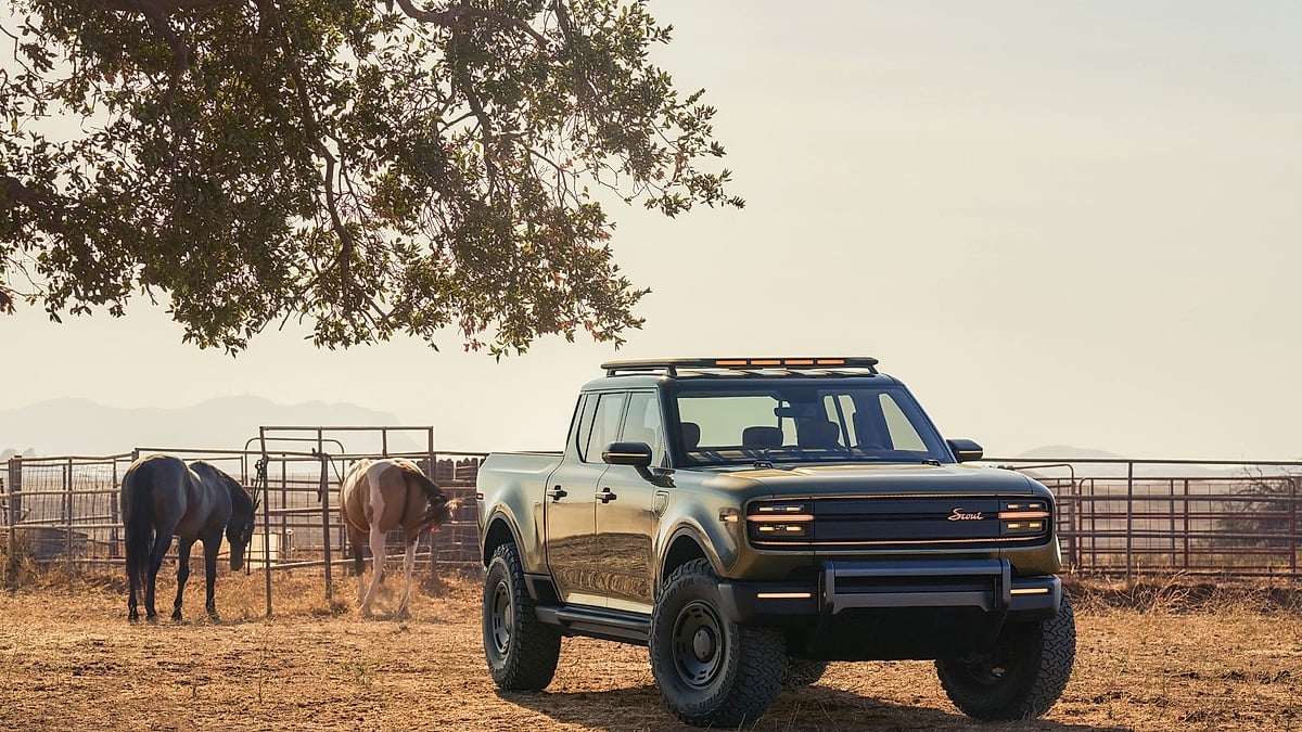 Scout Terra electric pickup truck in olive green at a ranch with horses in the background, showing the amber roof bar lighting and rugged off-road stance