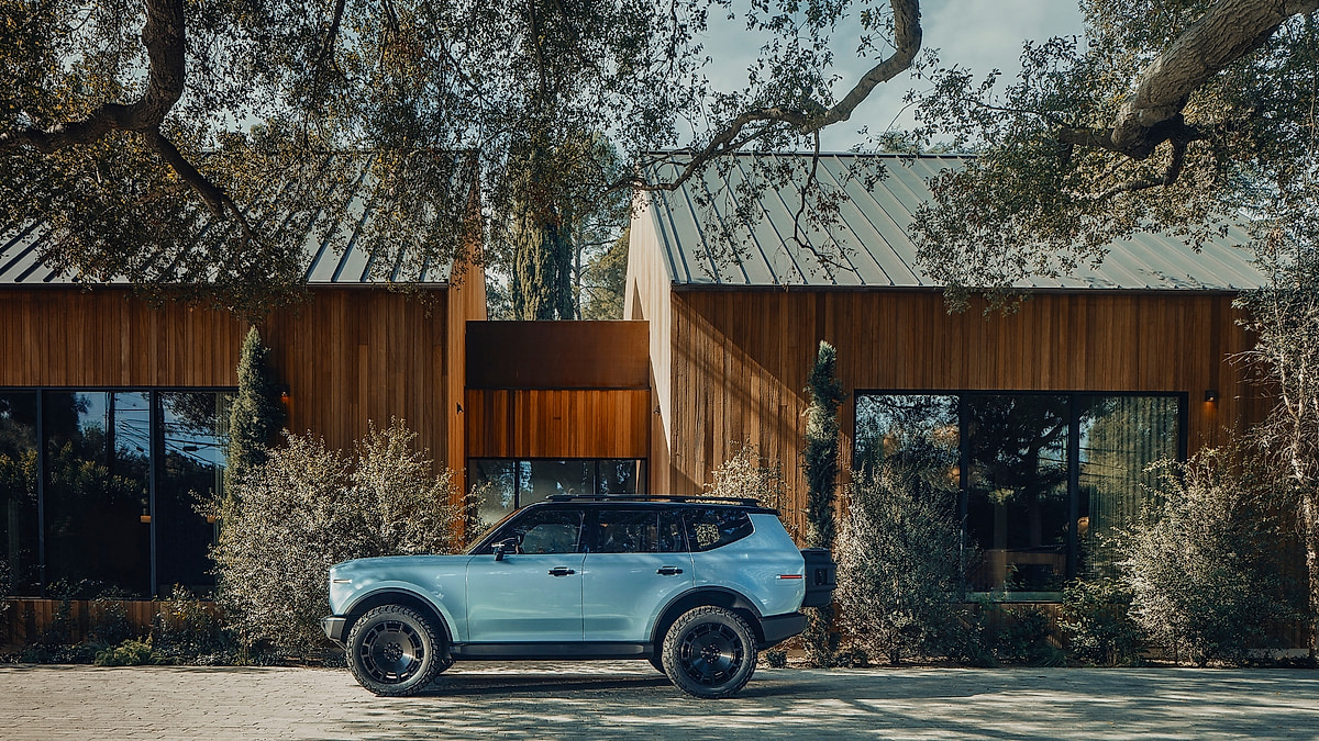 Side profile of the Scout Traveler electric SUV in light blue-grey parked in front of a contemporary wood-paneled architectural home surrounded by trees