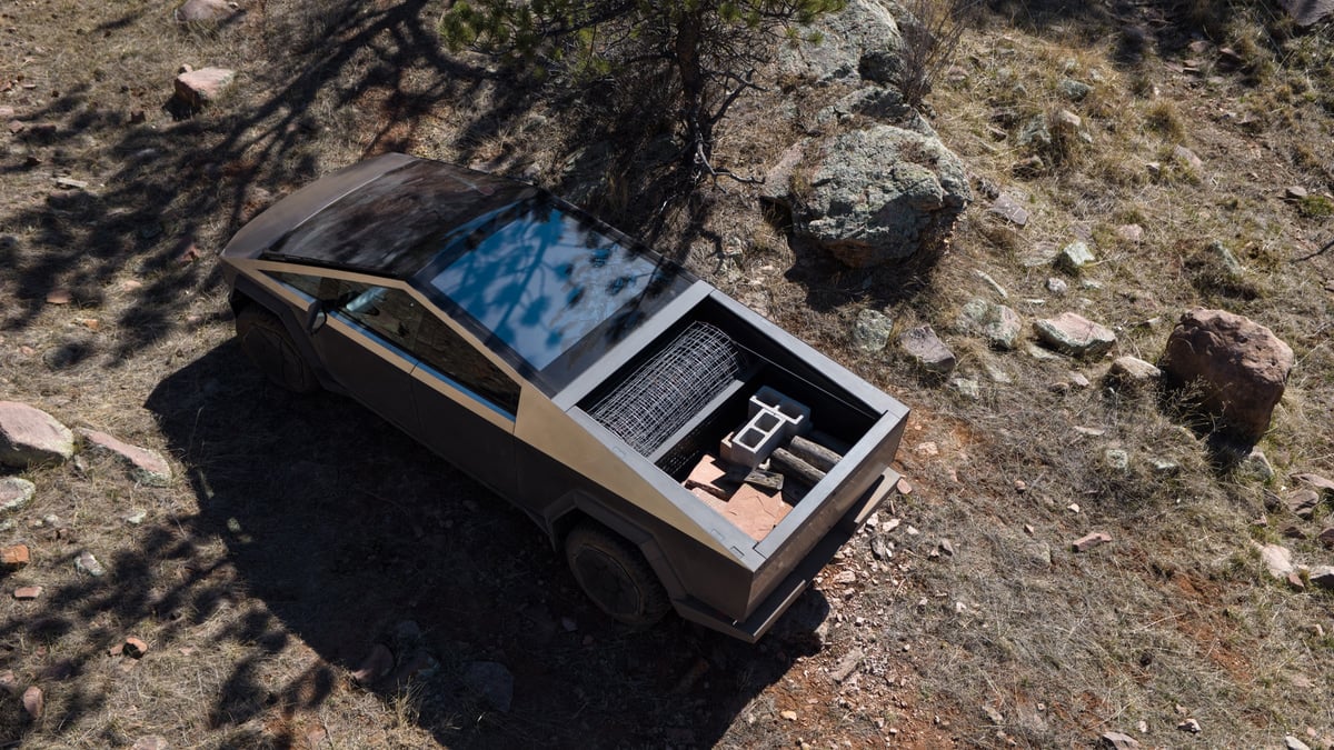 Aerial view of a Tesla Cybertruck parked on rocky terrain, bed loaded with wire fencing, cinder blocks, and firewood