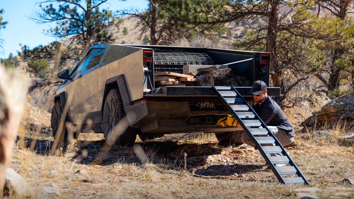 Tesla Cybertruck with its bed loaded with firewood and fencing material, a man setting up a loading ramp at the tailgate in a forested area
