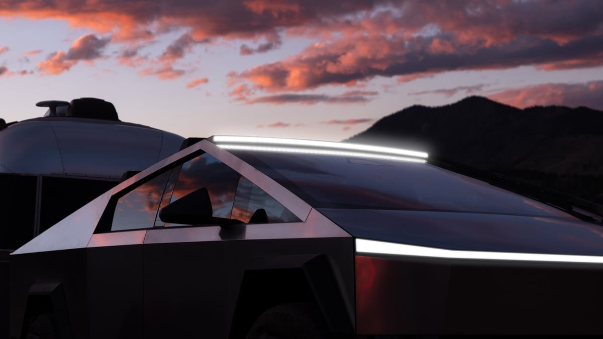 Close-up of a Tesla Cybertruck's angular roofline and light bar at sunset, towing a silver camper trailer with mountains in the background