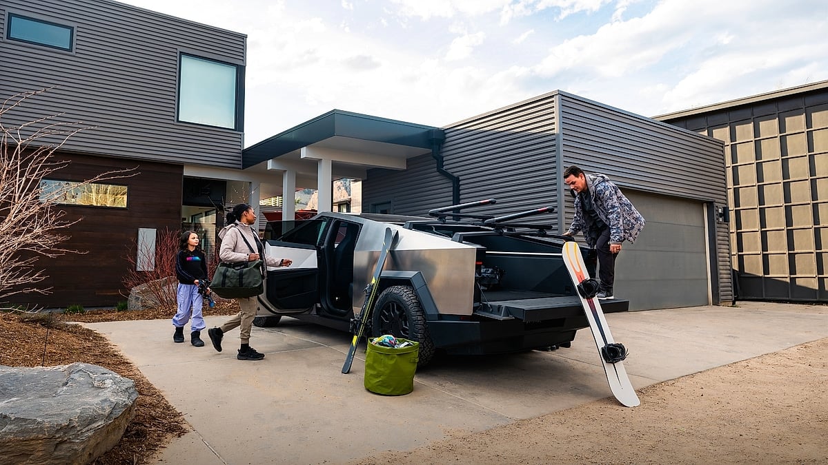 Tesla Cybertruck parked in a modern home driveway while people load snowboarding gear into the rear cargo area.