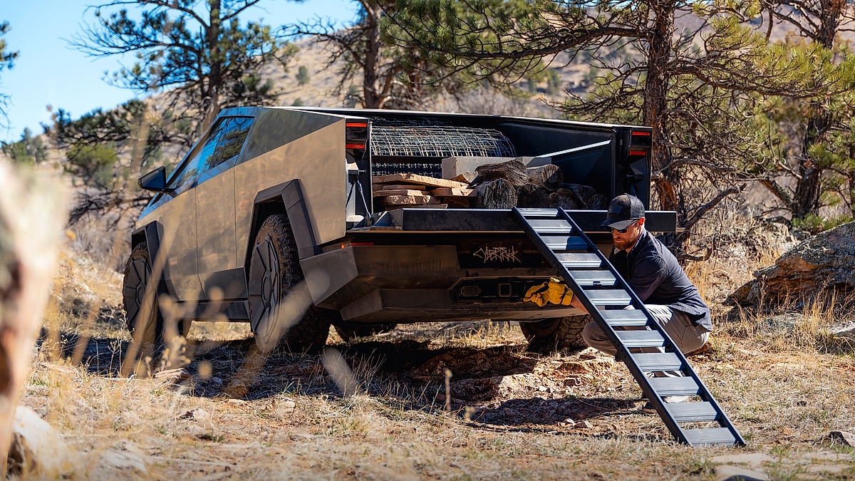 Tesla Cybertruck parked off-road with firewood in the bed and a ramp extended near a campsite.
