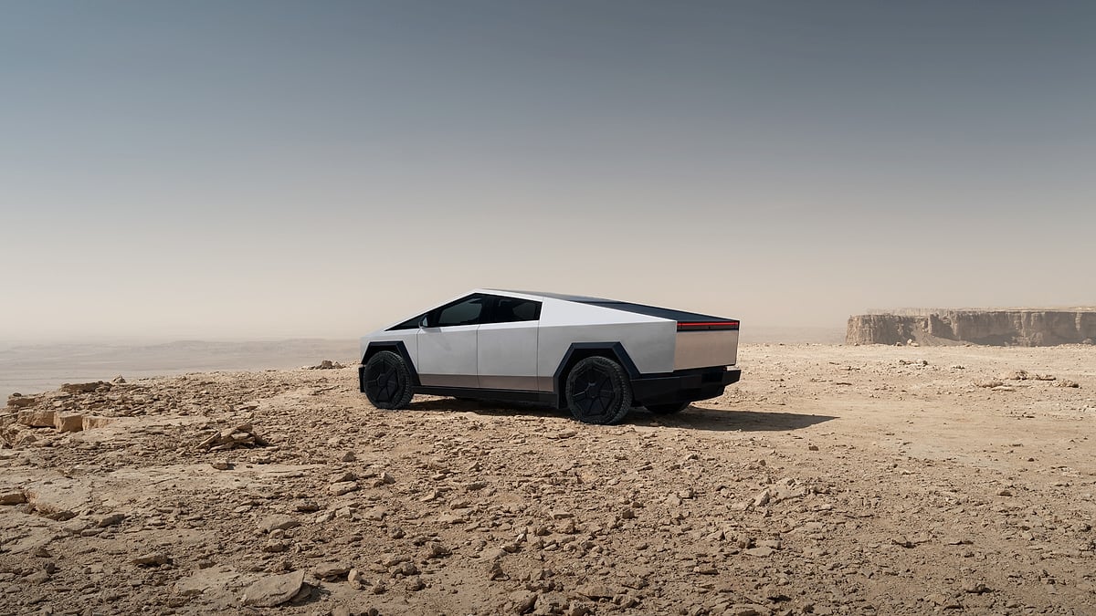 Tesla Cybertruck parked on a rocky desert plateau with cliffs in the distance under a clear sky.