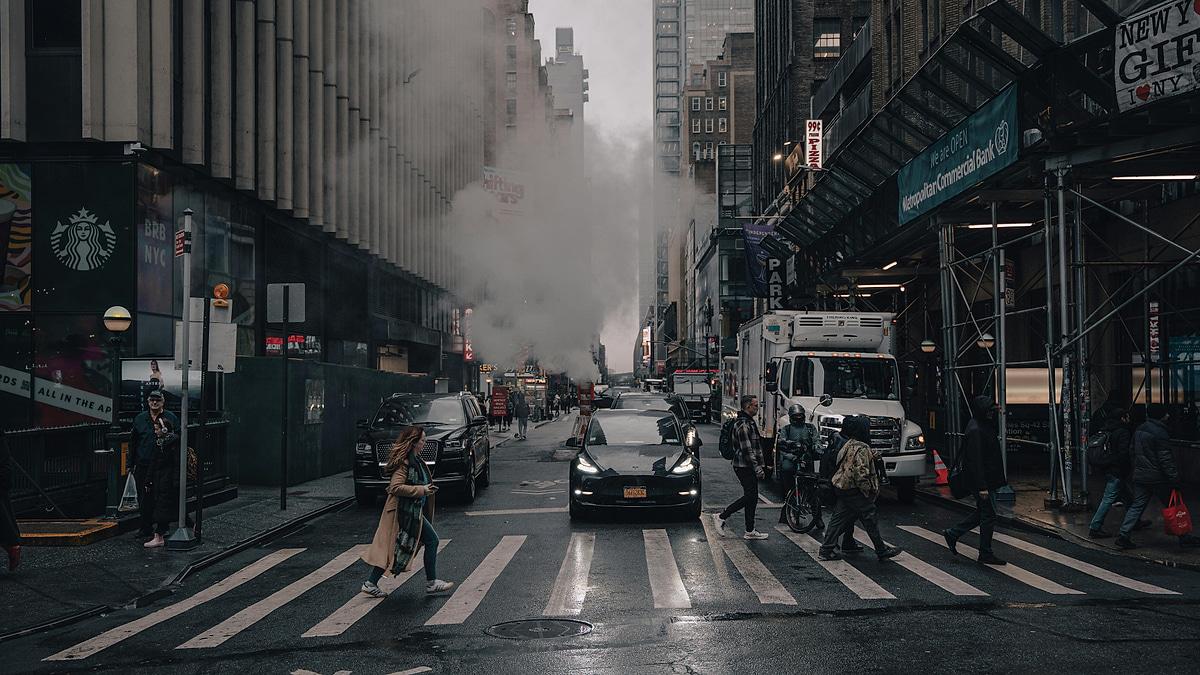 Black Tesla driving through a New York City crosswalk with street steam and pedestrians.