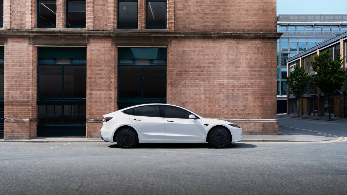 White Tesla Model 3 parked on a city street in front of a brick building, showing clean side profile and flush door handles.