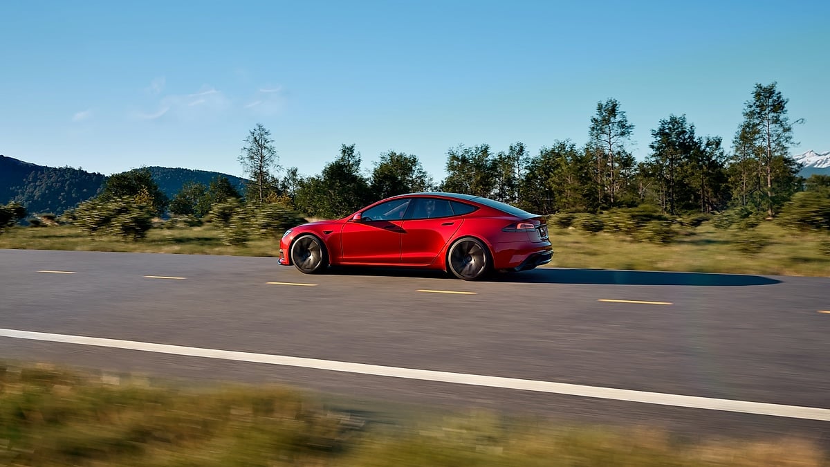 Side profile of a red Tesla Model S driving on an open road with trees and mountains in the distance.