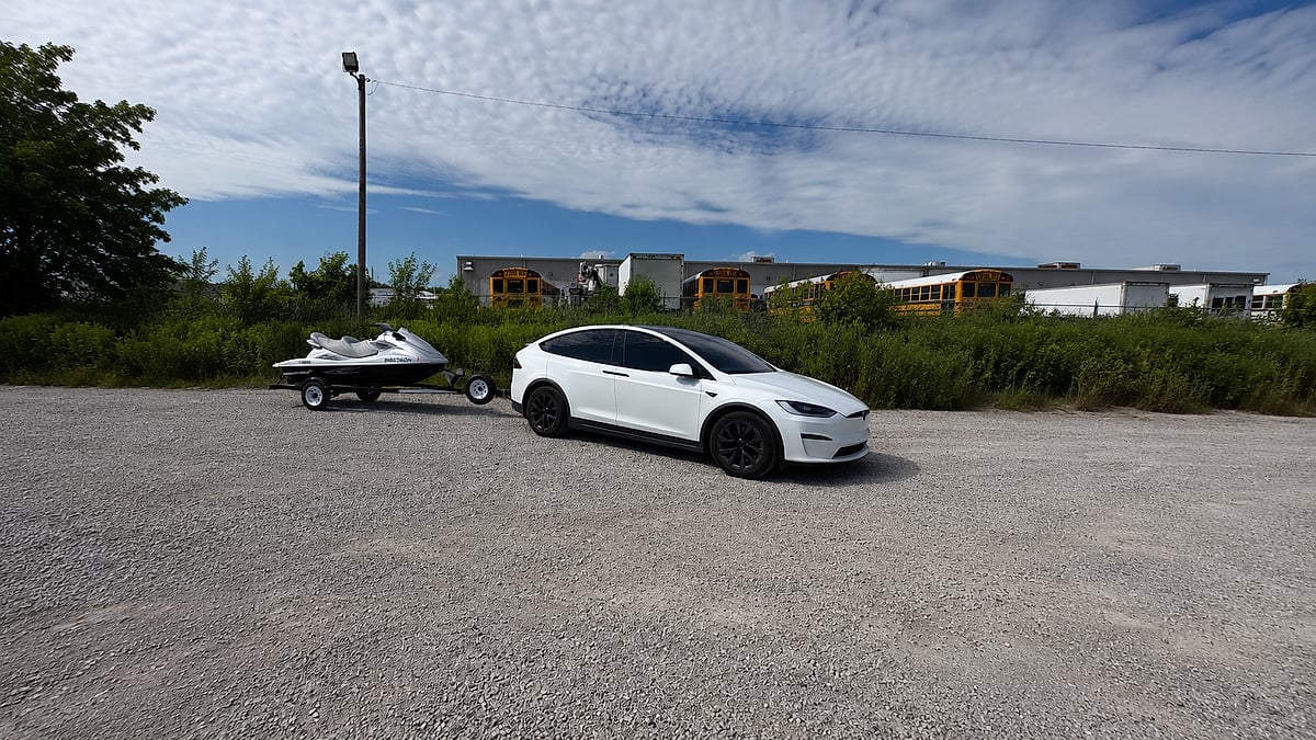 White Tesla Model X towing a personal watercraft on a small trailer in a gravel lot near school buses.
