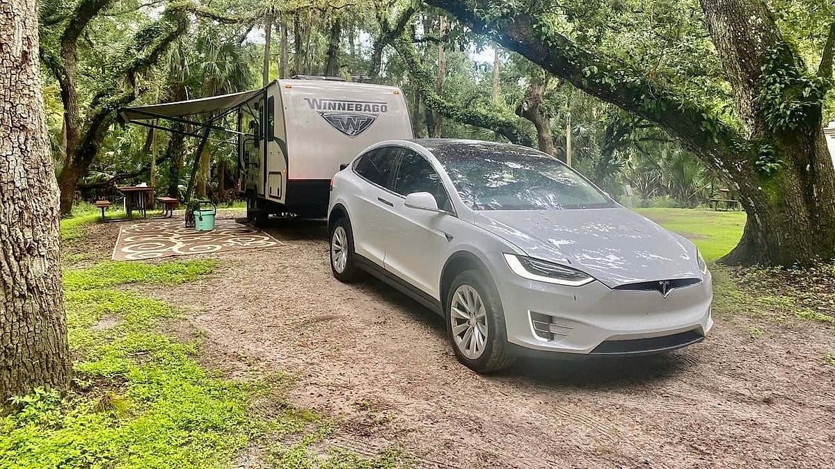 Tesla Model X parked at a wooded campsite while towing a Winnebago camper trailer.