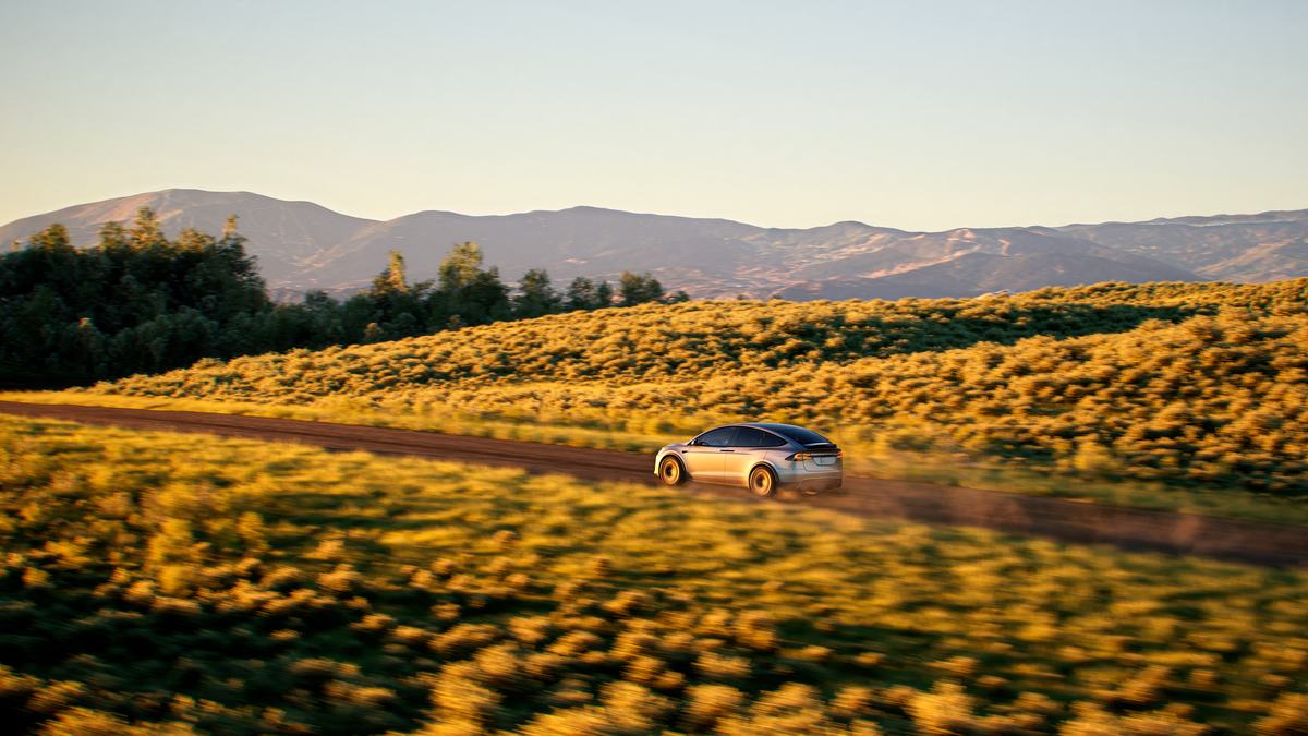 Tesla Model X driving on a rural dirt road through rolling green hills at sunset