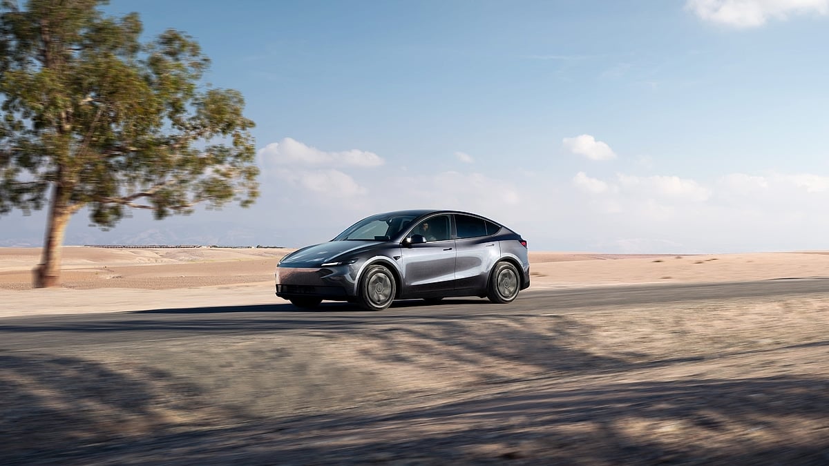 Refreshed Tesla Model Y in gray driving on desert road with sand dunes and lone tree in background, side profile view