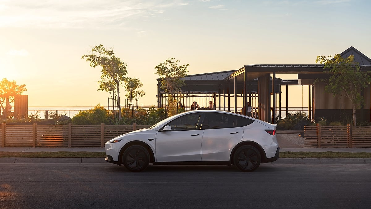 White Tesla Model Y shown in side profile on a road at sunset, with a waterfront building and warm evening light in the background.