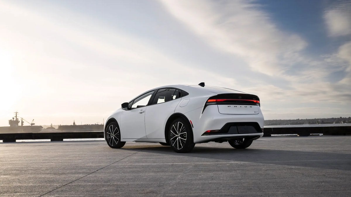 White Toyota Prius parked on a waterfront pier at sunset, rear three-quarter view emphasizing modern hybrid sedan design.