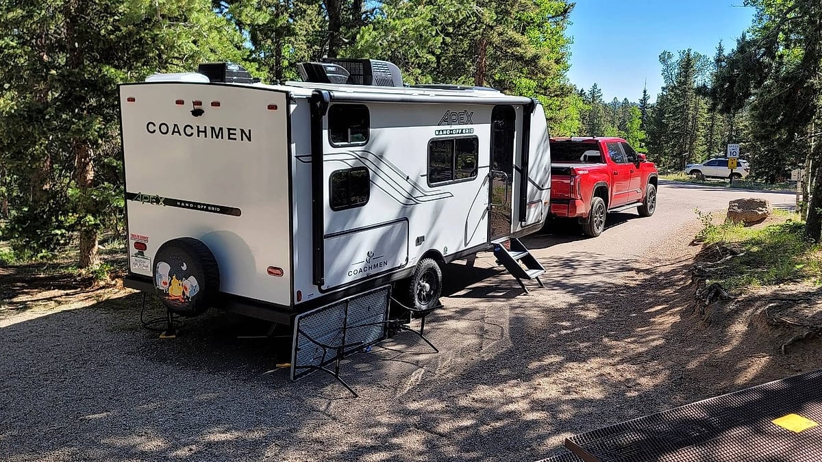 Red Toyota Tundra towing a Coachmen Apex camper trailer at a forest campsite.