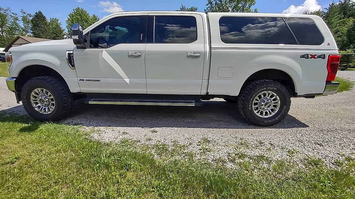 White Ford Super Duty crew cab pickup with a camper shell parked on gravel.