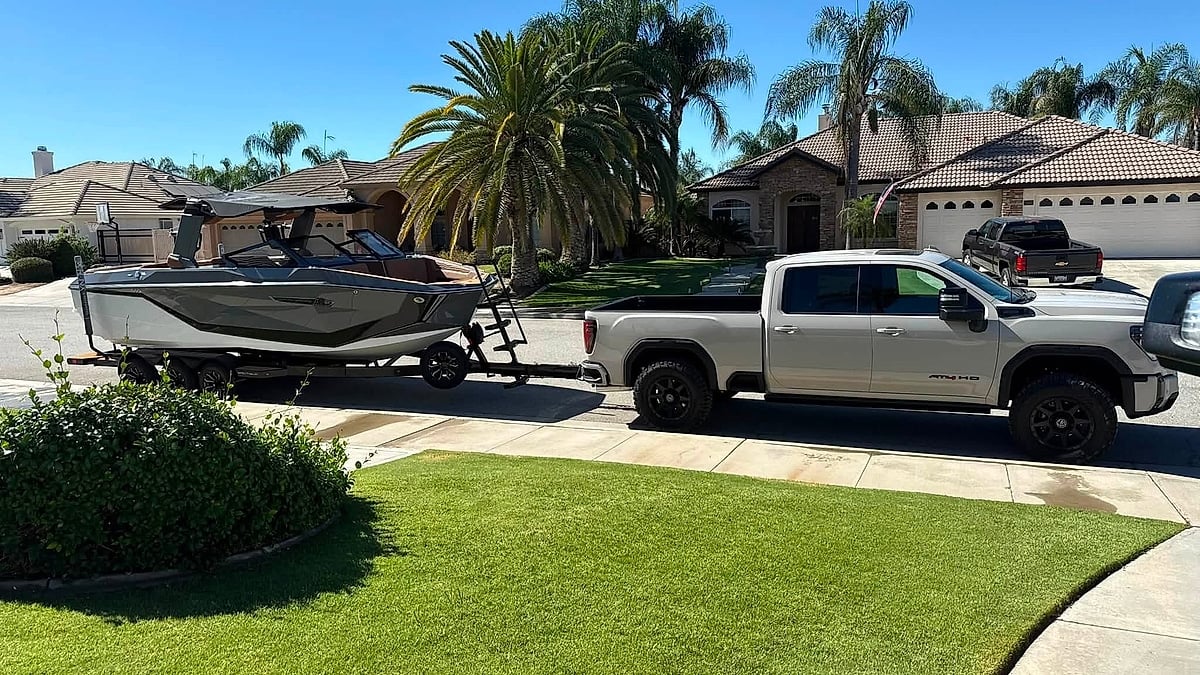 White GMC Sierra HD AT4 towing a covered MasterCraft boat on a sunny suburban street lined with palm trees.