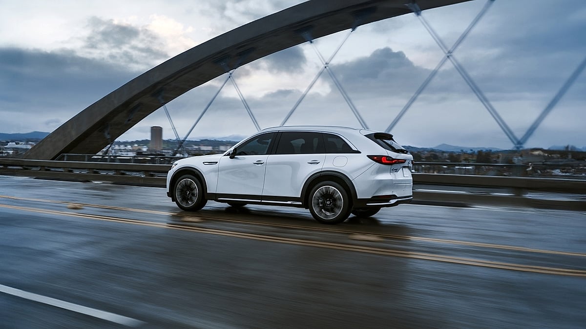 White Mazda CX-90 driving across a wet bridge under cloudy skies.