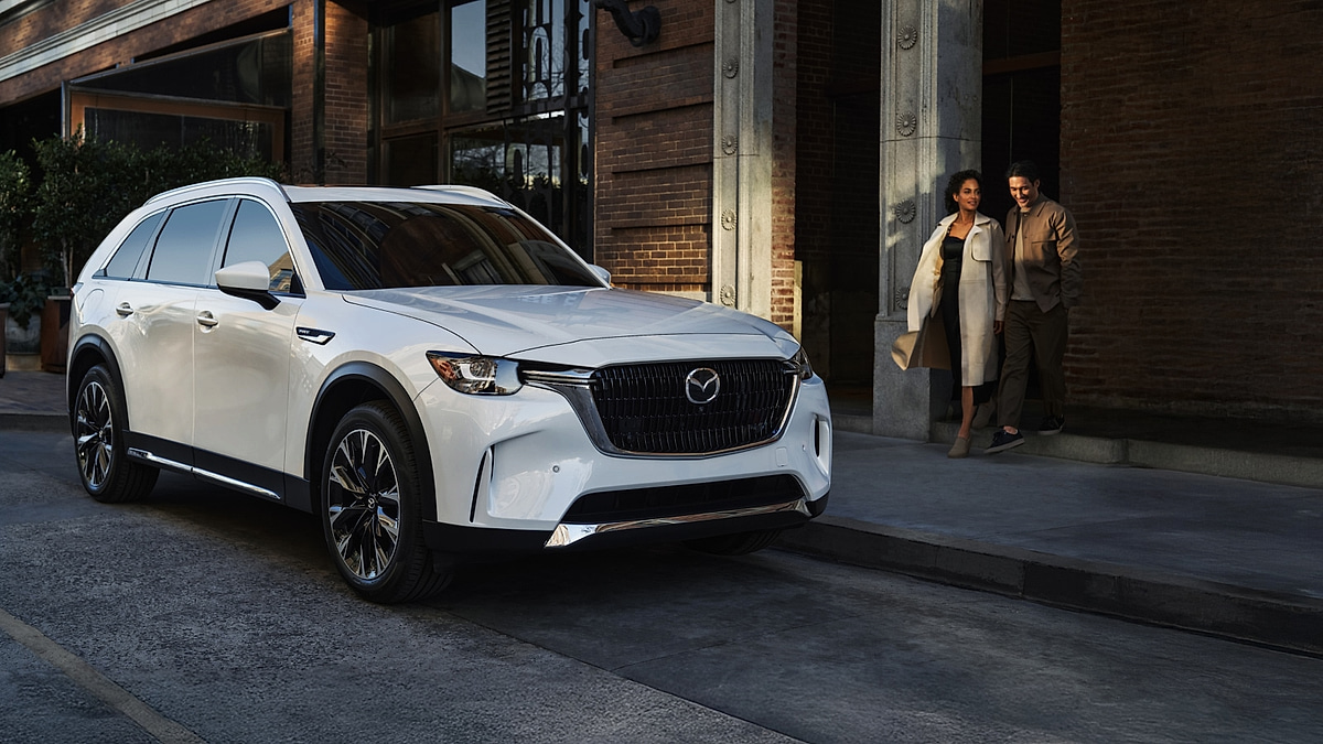White Mazda CX-90 parked on a city street outside a building entrance at dusk.