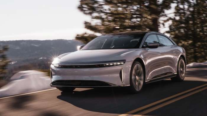 A white Lucid Air sedan is pictured driving on a mountain road with rolling hills in the background.