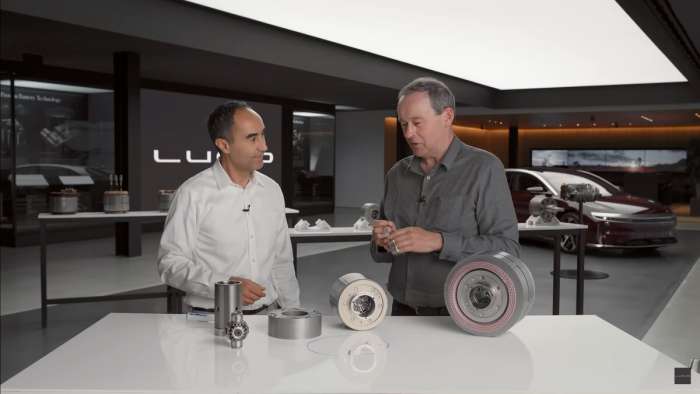 CEO Peter Rawlinson and Powertrain VP Emad Dlala explain the motor's design for Lucid's Tech Talk series. A Lucid Air is visible in the background while the motor is on a table in front of the men in various states of disassembly.