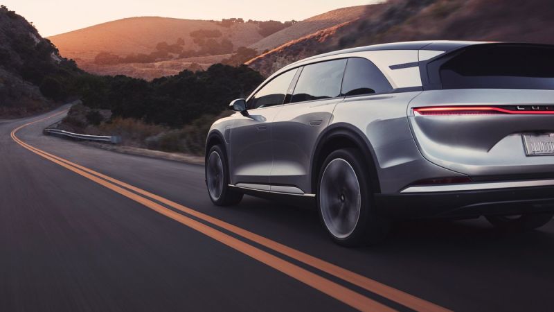 A sleek silver Lucid Gravity SUV drives along a winding road at sunset, surrounded by hills and lush greenery.