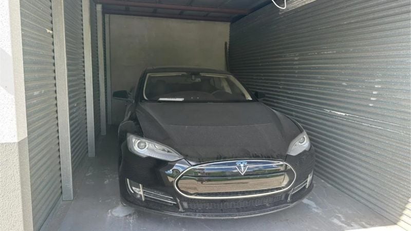 Black car with a covered hood parked in a narrow, corrugated metal garage.