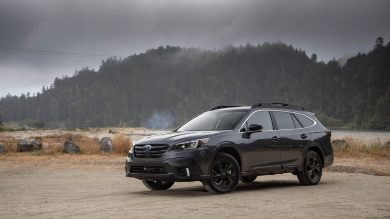 A dark gray 2021 Subaru Outback is shown from a front three-quarter view, parked on a gravel area with misty mountains and forest in the background.