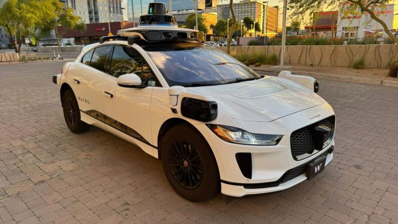 White Jaguar i-Pace with Waymo autonomous driving sensors, parked on brick pavement with urban backdrop