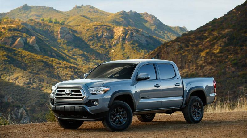 Front three-quarter view of a silver Toyota Tacoma off-road truck parked on a mountain overlook with rugged terrain behind it.