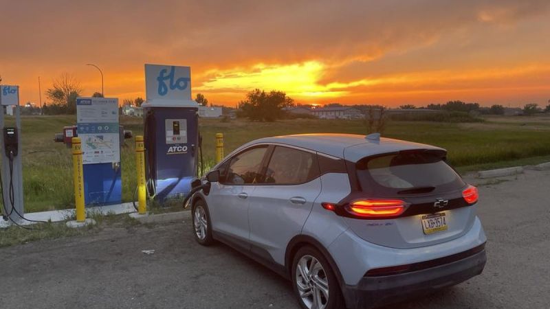 A Chevrolet Bolt EV charges at a Flo station under a vibrant sunset, with lush greenery in the background.