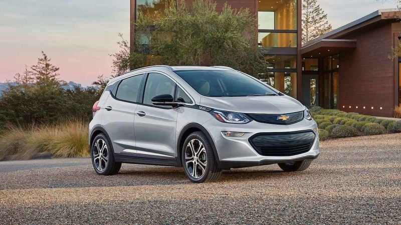 A silver 2022 Chevrolet Bolt EV parked on gravel, with a modern home and landscaped greenery in the background.