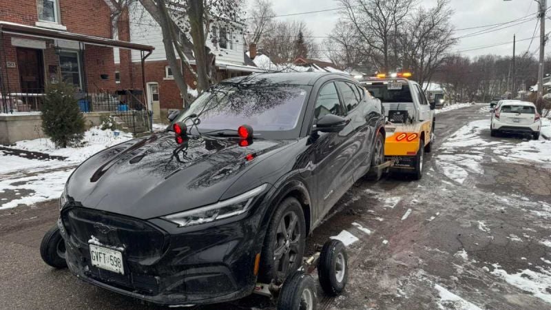 A black Ford Mustang Mach-E electric SUV, likely a 2022 model, is shown being loaded onto a tow truck from its front three-quarter angle on a snowy residential street.
