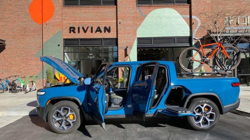 Electric blue Rivian R1T pickup truck with open doors, parked in front of Rivian headquarters, with orange bicycle mounted on roof rack.
