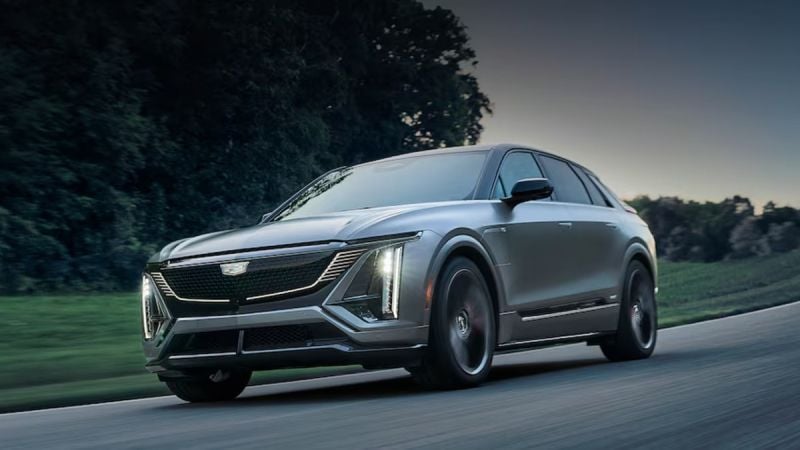 2023 Cadillac Lyriq electric SUV driving on a road, front three-quarter view showing illuminated LED grille and vertical headlights.
