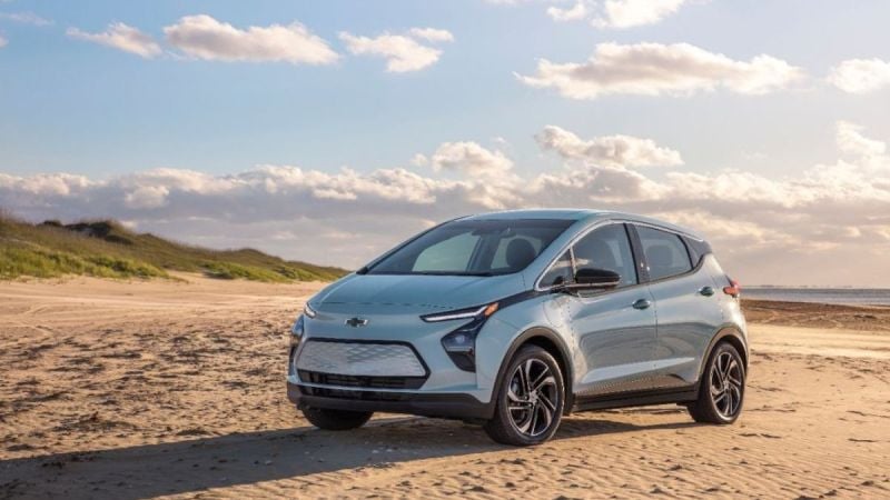A silver Chevrolet Bolt EV is parked on a sandy beach under a clear blue sky with scattered clouds. The scene conveys a serene and eco-friendly vibe.