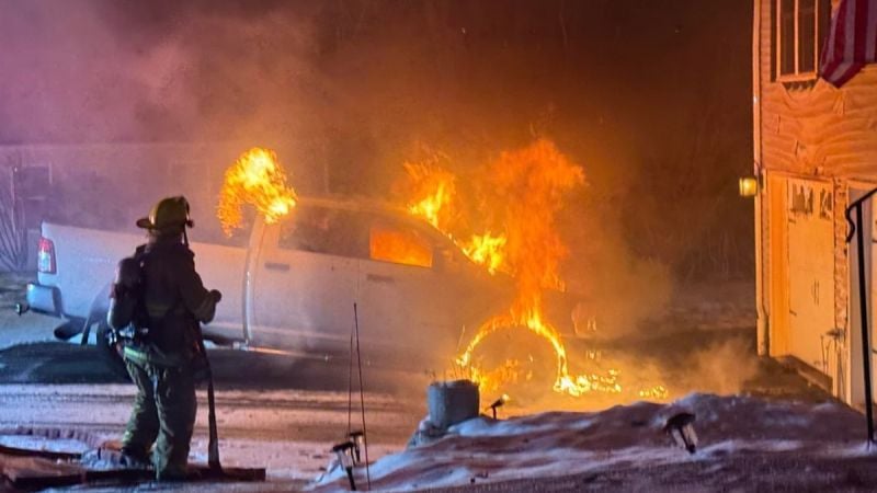 A firefighter sprays water on a white Ram truck that is on fire in a driveway at night.