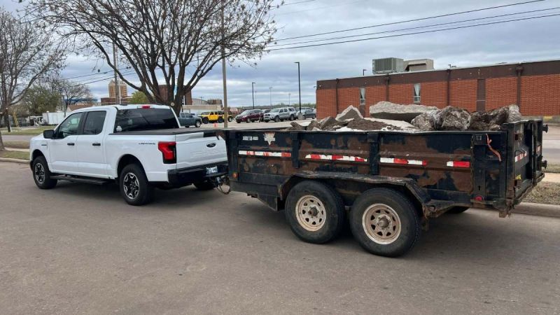 A white 2023 Ford F-150 Lightning pickup truck, likely a recent model year, is shown from the rear quarter view while connected to a dark-colored dual-axle trailer loaded with construction debris.