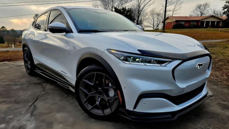 2023 Ford Mustang Mach-E in Star White Metallic parked on a suburban driveway, front three-quarter low angle view with a brick house and bare trees in the background.