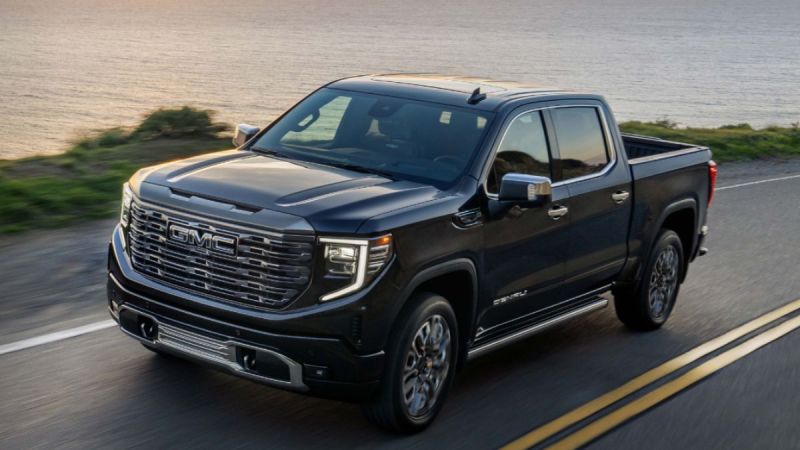 A black 2023 GMC Sierra 1500 Denali truck drives on a coastal road at sunset, with ocean and greenery in the background. The scene conveys power and adventure.